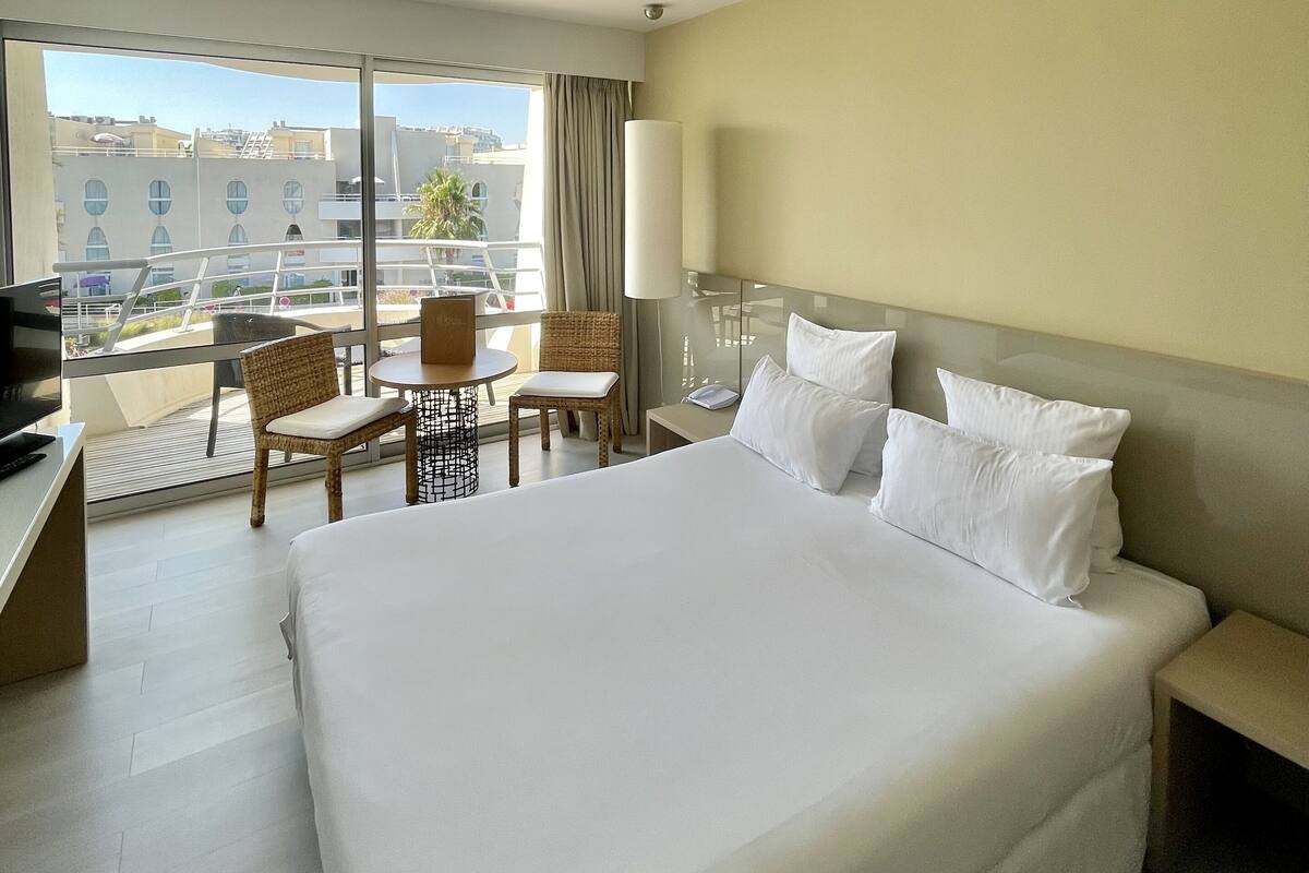 Hotelkamer met een groot bed, een bureau, een stoel, een lamp en uitzicht op de stad door grote ramen.