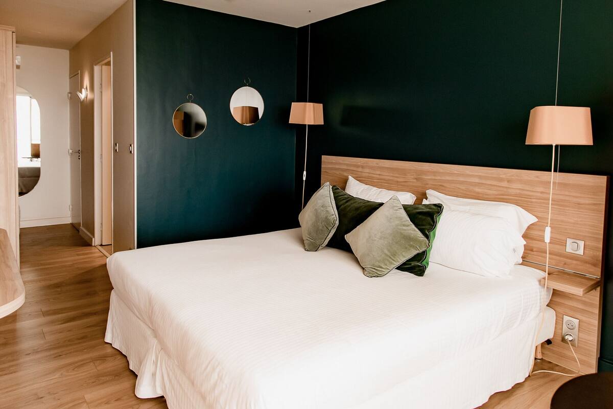 Een netjes opgemaakt bed met witte lakens en meerdere kussens, een houten hoofdbord en twee wandlampen.
