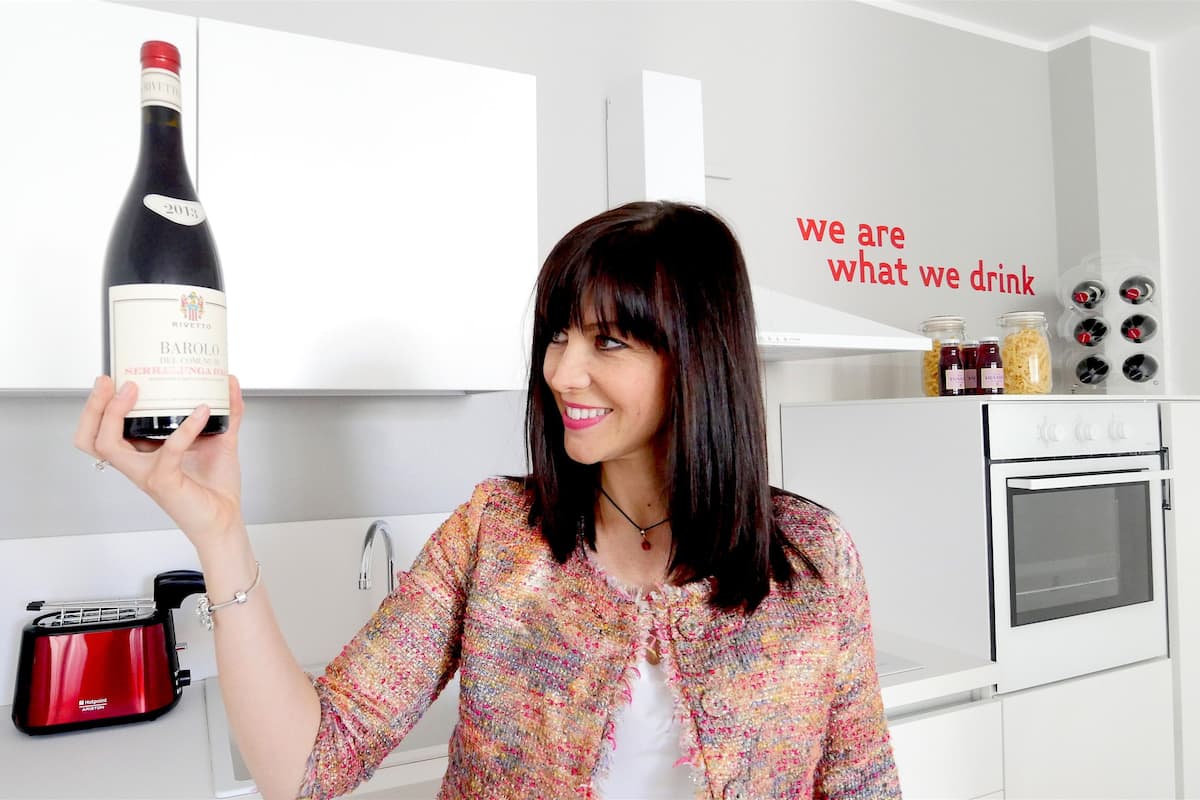 A person holding a bottle of wine in a modern kitchen with white cabinets, a red coffee maker, and a wine rack.
