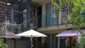 A courtyard with a grassy area, a concrete planter with purple flowers, a white umbrella, and a seating area with a purple chair. - Rivetto Suites (Alba)