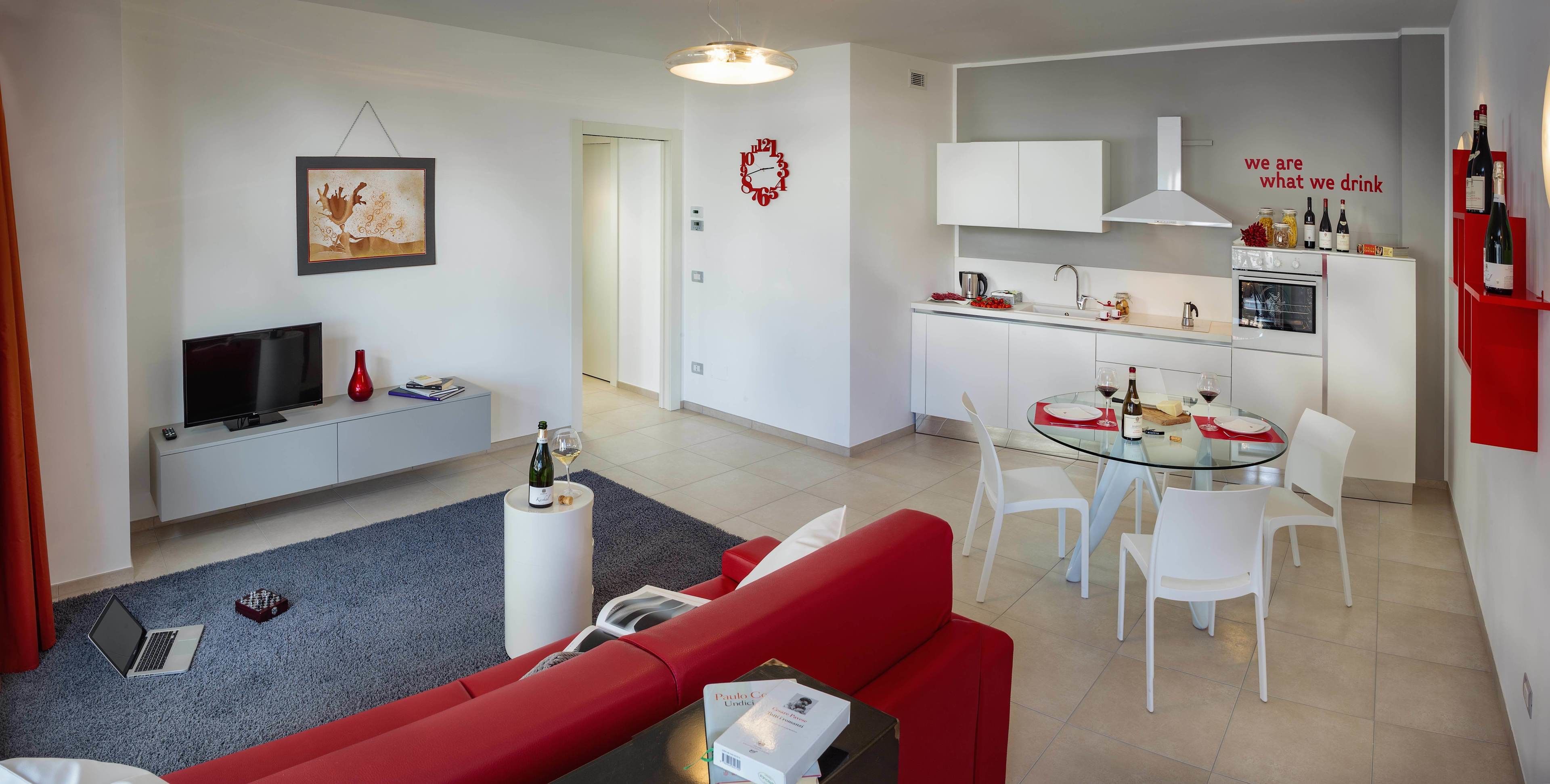 A modern living room with a red sofa, a glass coffee table, and a TV. There's a kitchen with white cabinets, a dining table with white chairs, and a red wall with a wine rack.
