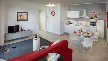 A modern living room with a red sofa, a glass coffee table, and a TV. There's a kitchen with white cabinets, a dining table with white chairs, and a red wall with a wine rack.