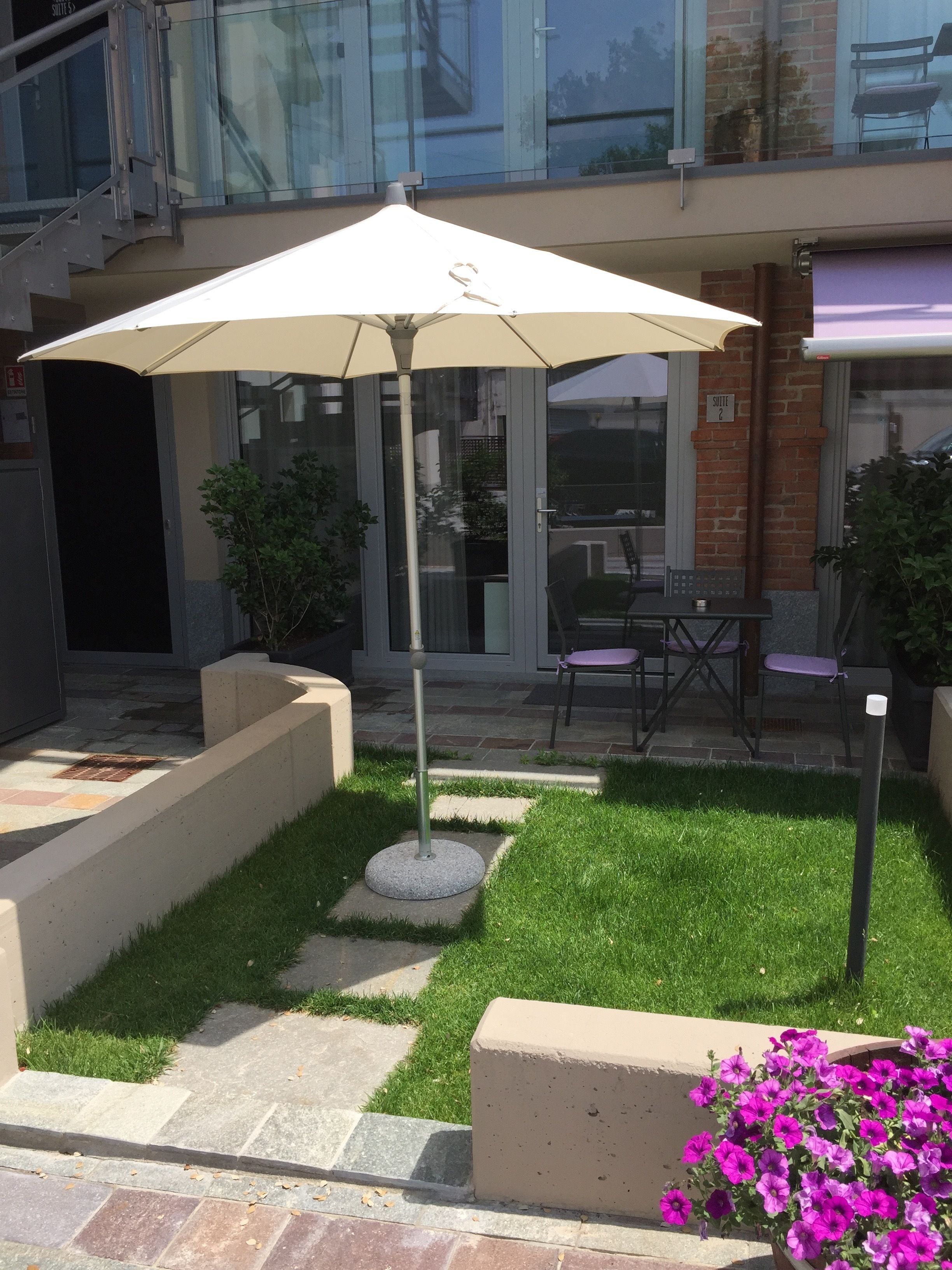 An outdoor patio area with a round table and chairs, a large umbrella, and a potted plant with purple flowers.