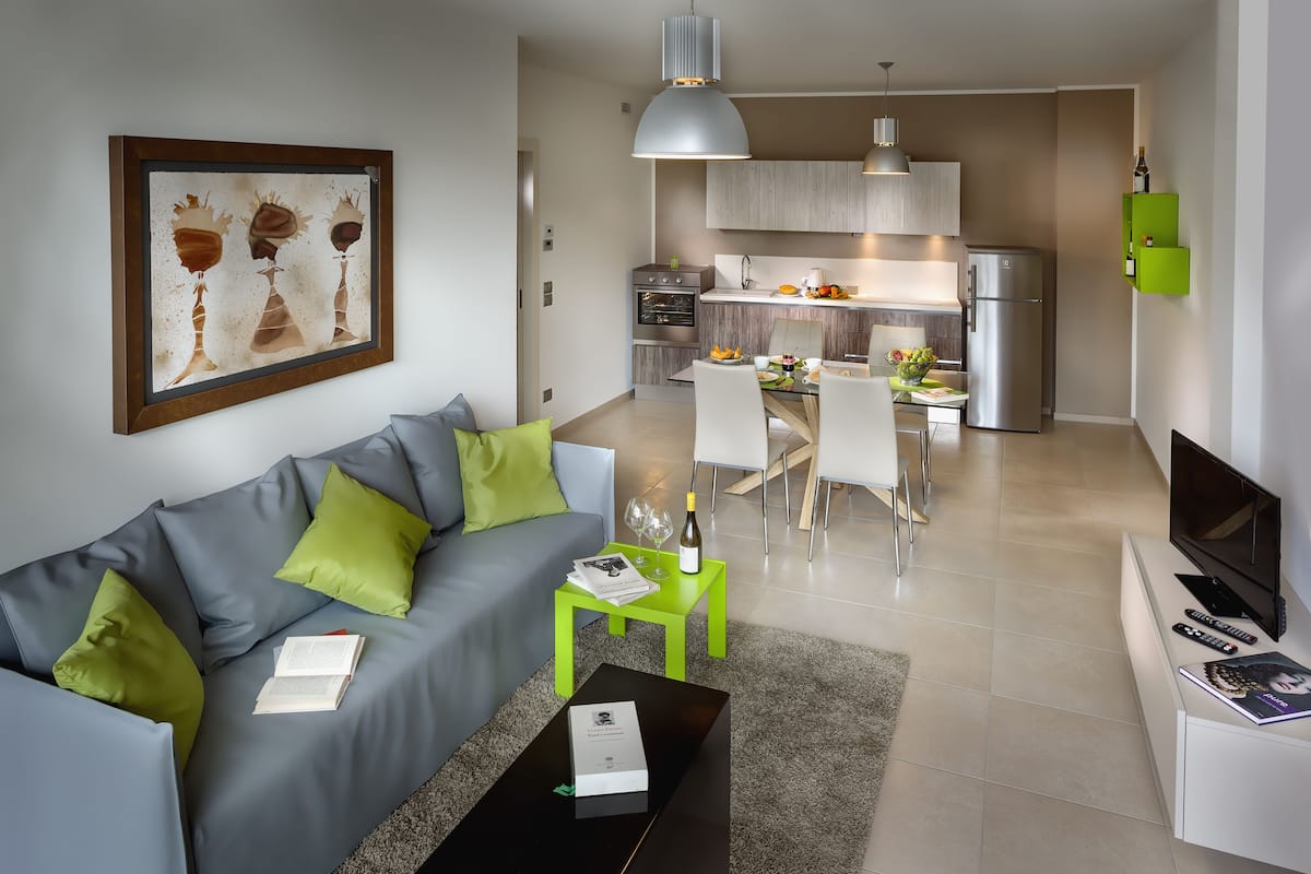 A modern living room with a grey sofa, green cushions, a coffee table with a book, and a TV on a stand.