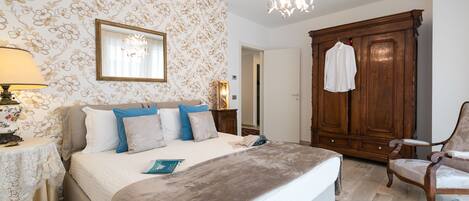 A bedroom with a bed, a wooden wardrobe, a chair, a table with a lamp, and a patterned wall.