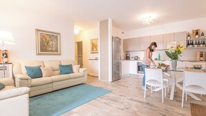 A modern living room with a beige sofa, a blue rug, and a kitchen area with white cabinets and a dining table. - Rivetto Suites (Alba)