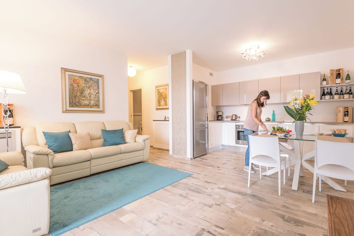 A modern living room with a beige sofa, a blue rug, and a kitchen area with white cabinets and a dining table.