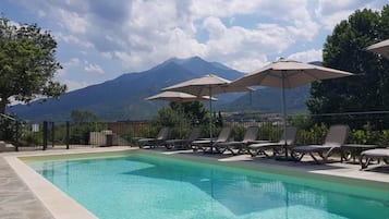 A swimming pool with lounge chairs and umbrellas, surrounded by a fence, with a mountain in the background.