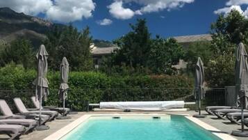 A clear blue swimming pool with lounge chairs and umbrellas, surrounded by greenery and a mountainous backdrop.