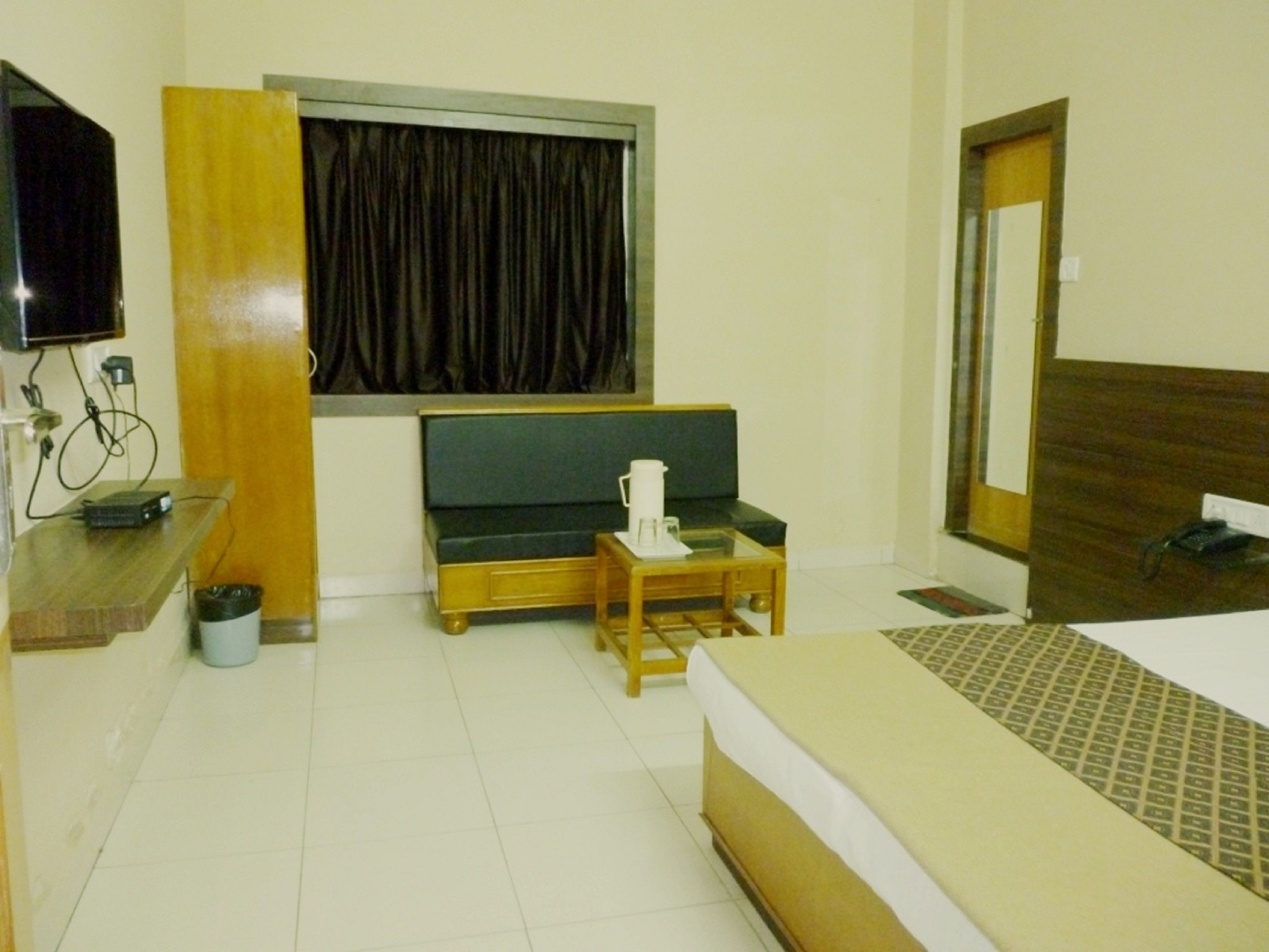 A hotel room with a bed, a small table with a cup, a television, and a window with curtains.