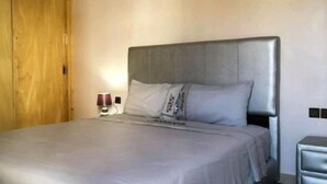 A bed with a grey headboard, a bedside table with a lamp, and a wooden wardrobe. - Soleil d'Agadir Centre (Agadir)