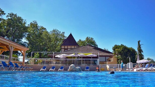 Une piscine avec des chaises longues et des parasols, un espace couvert doté d’un toit traditionnel, et un bâtiment avec un toit en forme de flèche en arrière-plan.