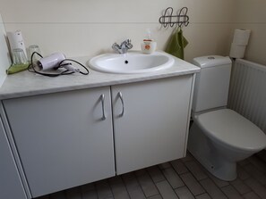 Une salle de bain avec un lavabo, des toilettes et un meuble de couleur blanche. Une serviette verte est accrochée au mur et un flacon de savon est posé sur le lavabo.