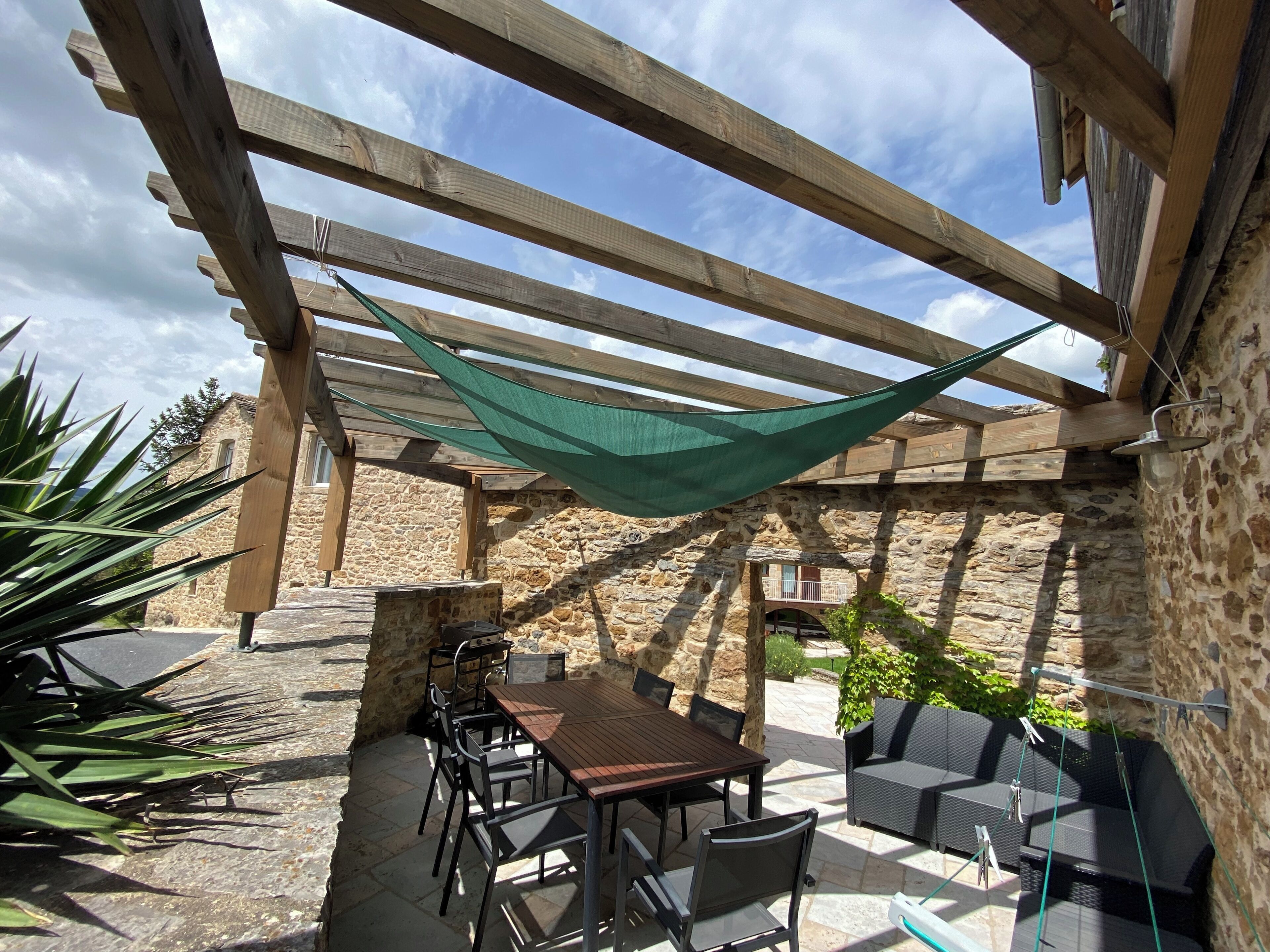 An outdoor patio with a wooden table and chairs, a hanging green shade, stone walls, and a stone building in the background.