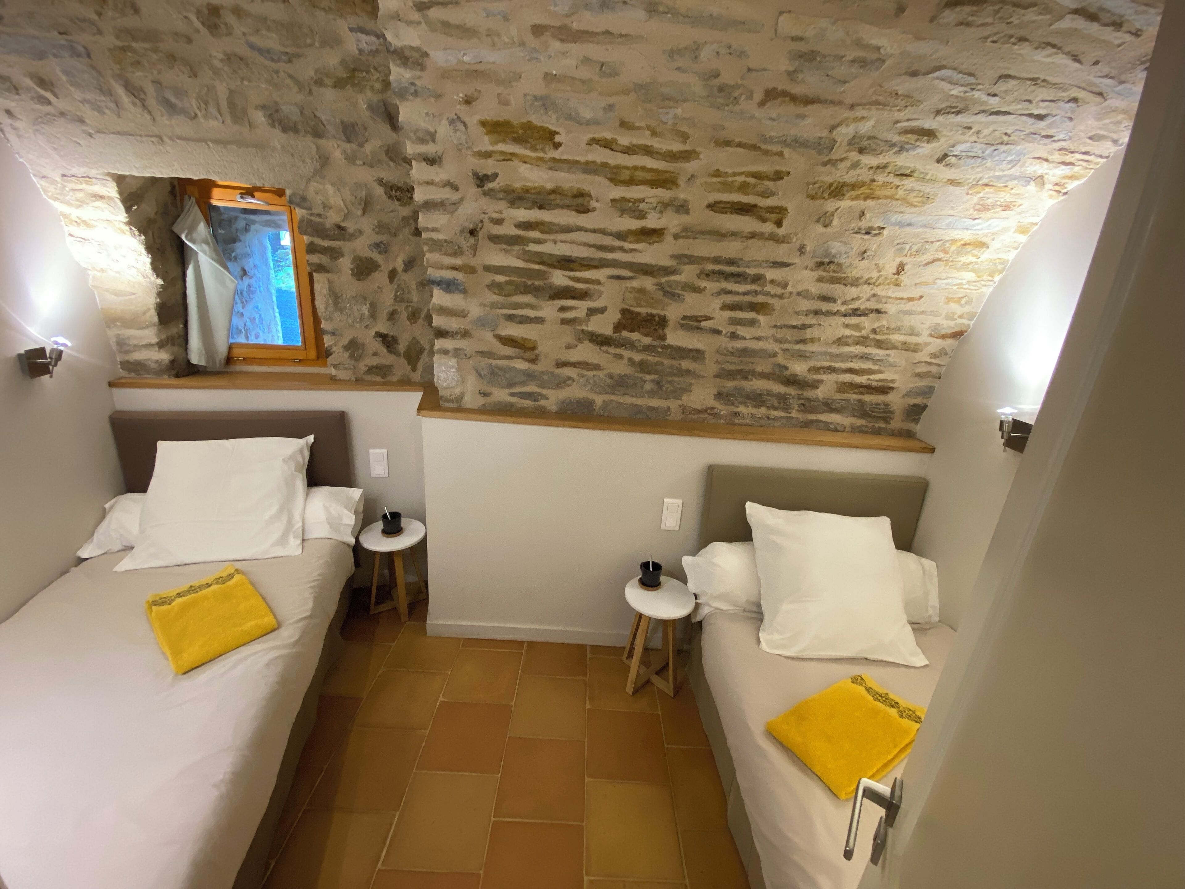 Une chambre avec deux lits, chacun étant équipé de draps blancs et d’une serviette jaune. Une petite table ronde se trouve entre les lits, et un mur en pierre se dresse au-dessus des lits.