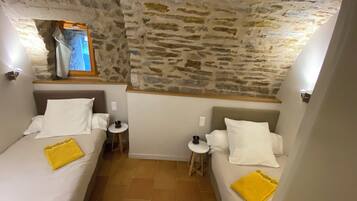 Une chambre avec deux lits, chacun étant équipé de draps blancs et d’une serviette jaune. Une petite table ronde se trouve entre les lits, et un mur en pierre se dresse au-dessus des lits.