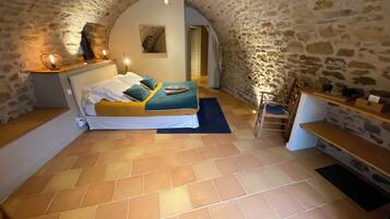 A cozy room with a stone wall, a bed with a blue and yellow bedspread, a wooden bench, and a small table with a lamp.