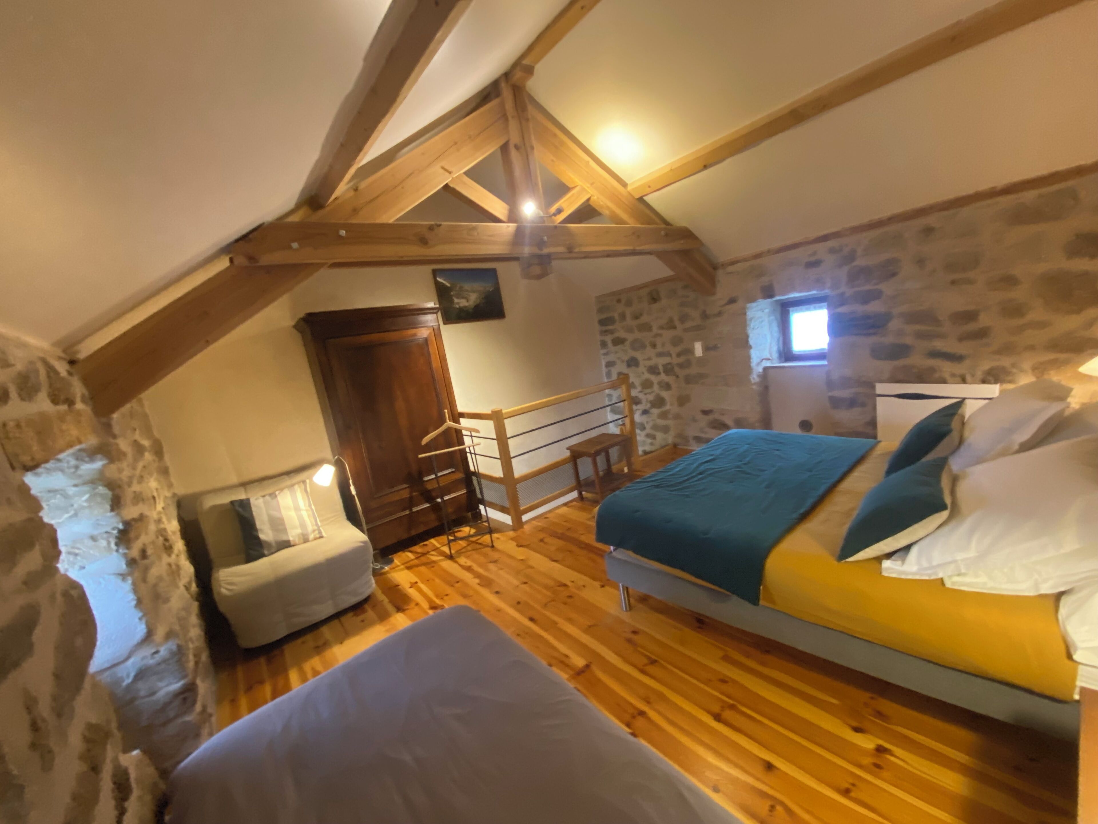 A cozy attic bedroom with a sloped ceiling, wooden beams, a stone wall, a bed with pillows, a small chair, and a wardrobe.