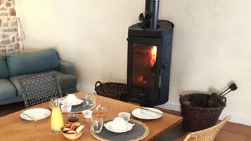 Et hyggeligt spiseområde med et bord dækket til morgenmad, en brændeovn, en fletstol og en blå sofa i baggrunden.