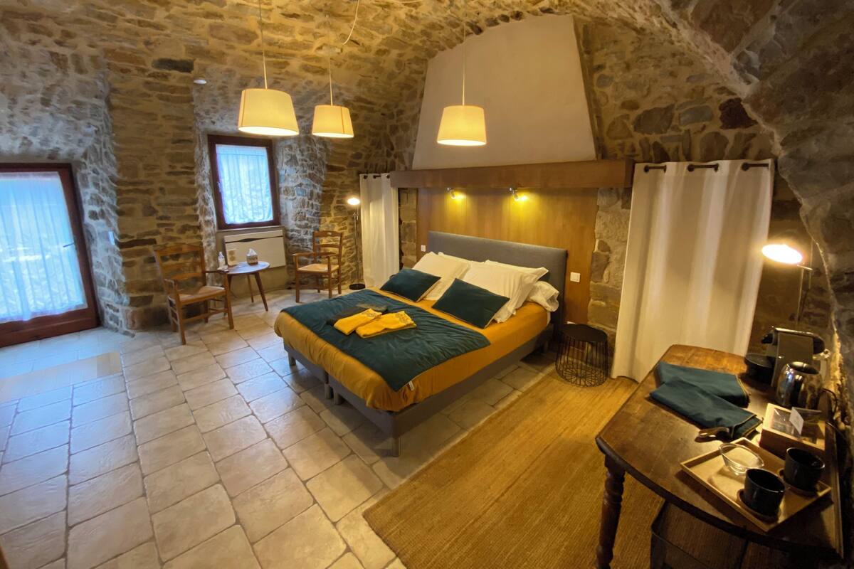 Quarto com paredes de pedra, uma cama, duas luminárias pendentes, uma mesa de cabeceira de madeira e uma janela com cortinas.