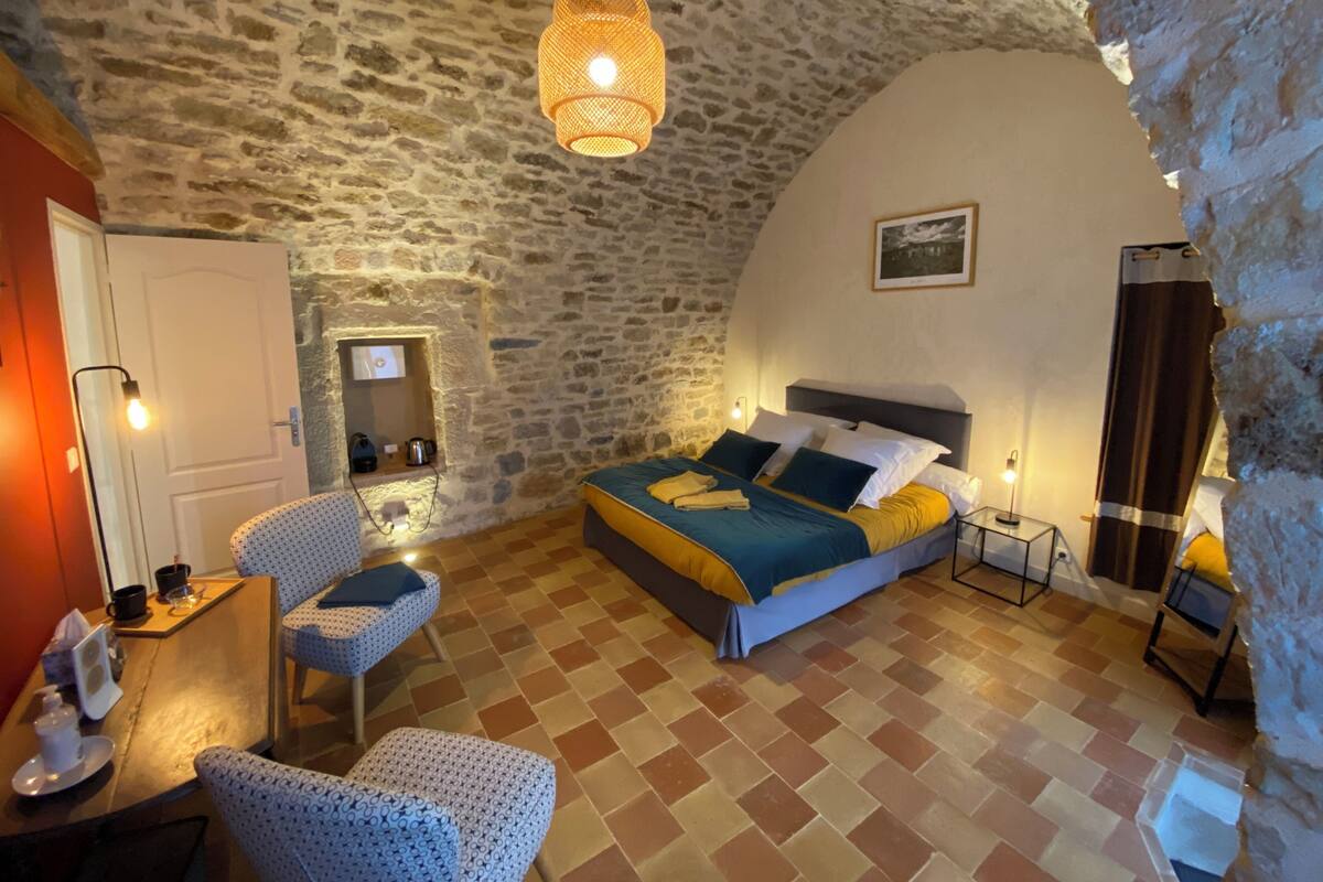 Quarto aconchegante com parede de pedra, cama com roupa de cama azul e amarela, uma cadeira, uma pequena mesa com luminária e um lustre.