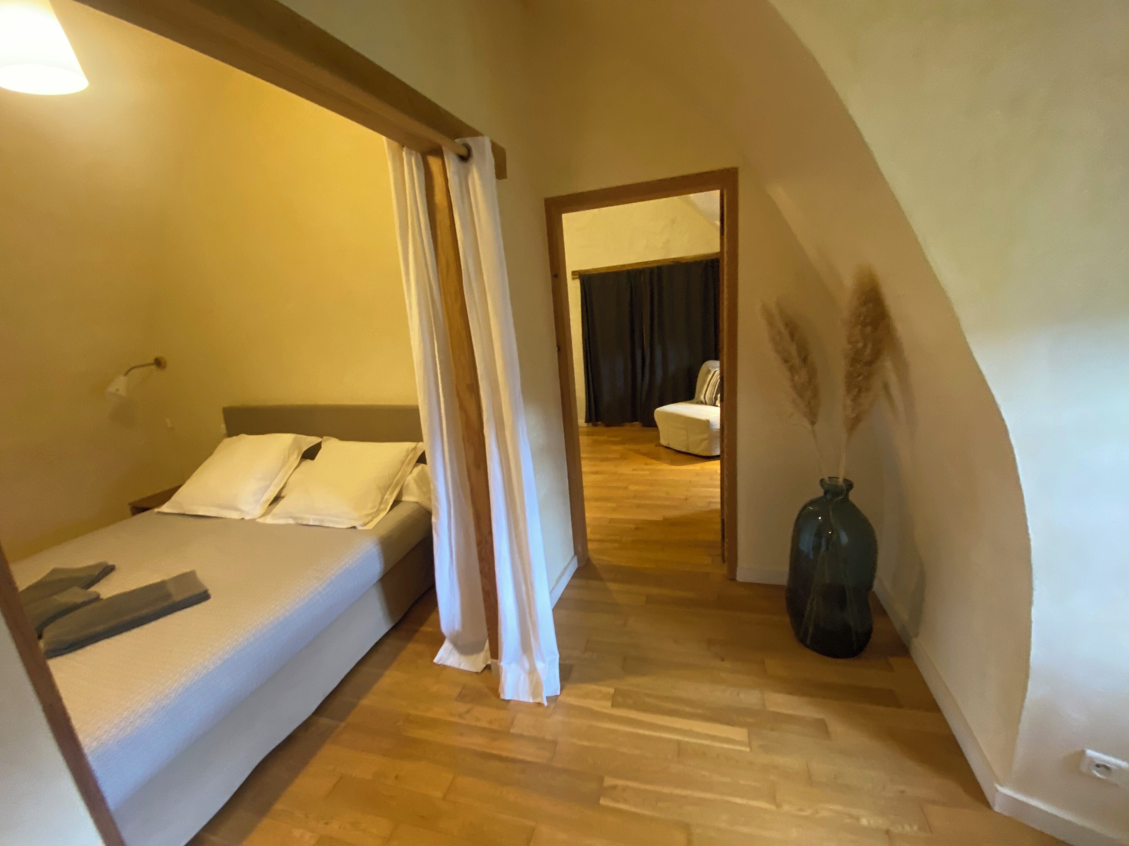 A bedroom with a bed, a wooden floor, a nightstand, a lamp, and a view of another room with a chair and a vase.