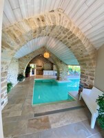 An indoor swimming pool with stone walls and a ceiling, surrounded by potted plants and a white bench.