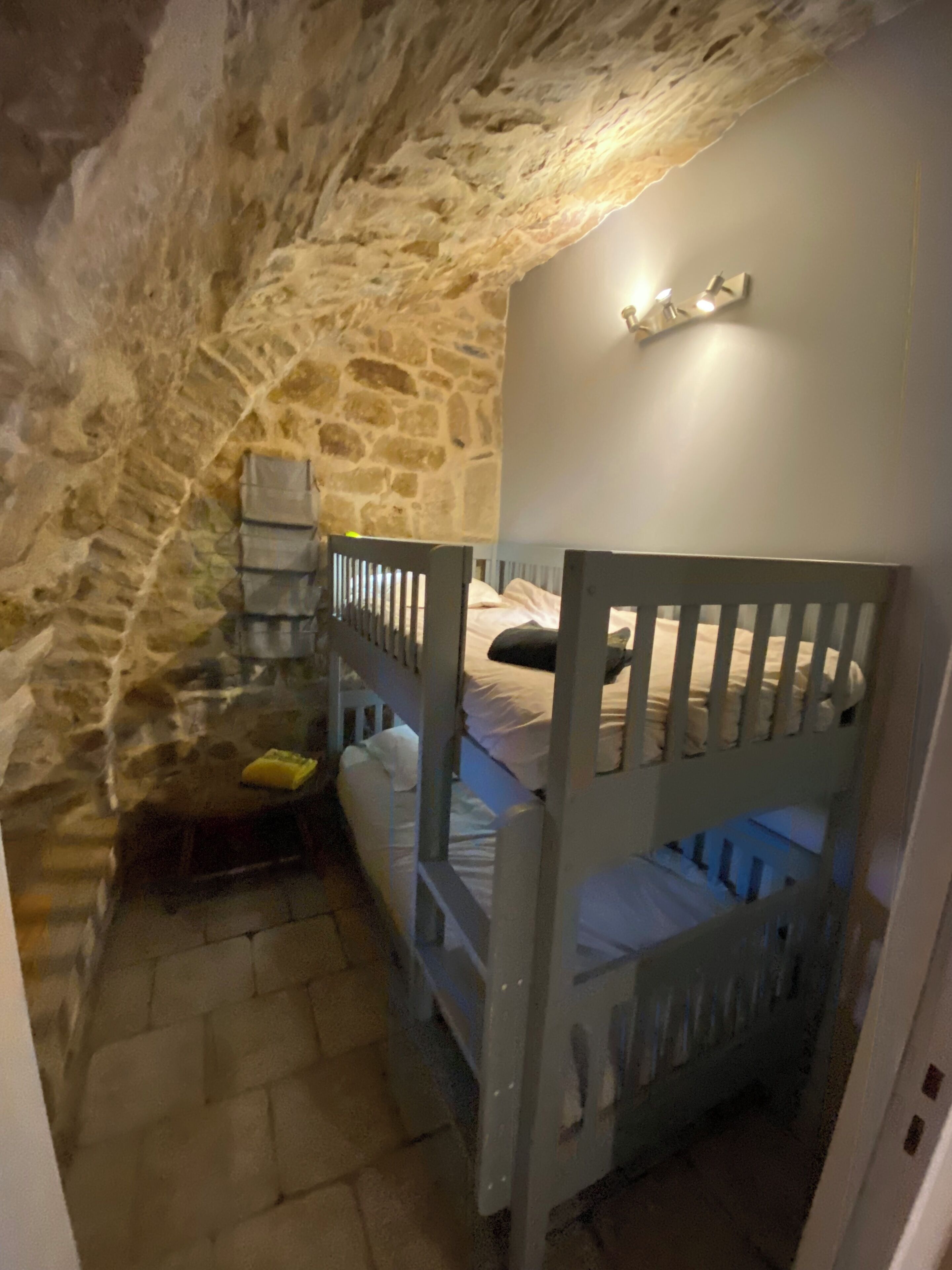 Una habitación con paredes de piedra, literas, una mesita y un aplique en la pared.