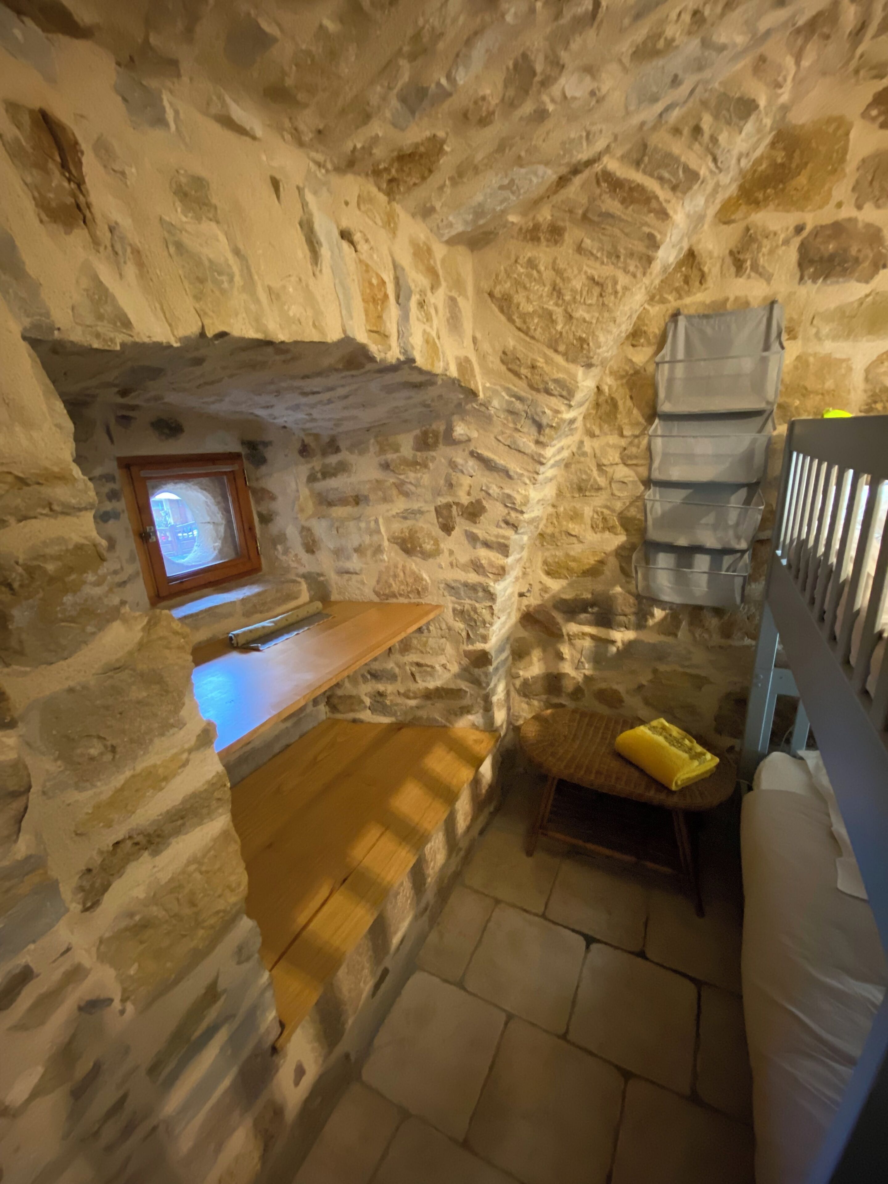 A stone-walled room with a wooden bench, a small window, and a bed with a mattress and pillows.