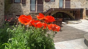 Un edificio de piedra con balcón, un puente de madera y amapolas rojas al frente.