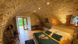 Un dormitorio acogedor con techo de piedra, una cama con ropa de cama verde y blanca, un escritorio con una silla y una ventana con vistas a un jardín.