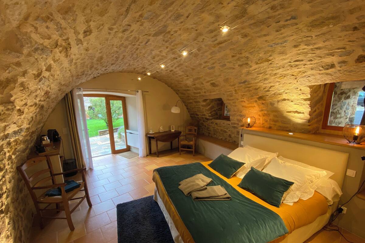 Um quarto aconchegante com teto de pedra, cama com roupa de cama verde e branca, escrivaninha com cadeira e janela com vista para um jardim.
