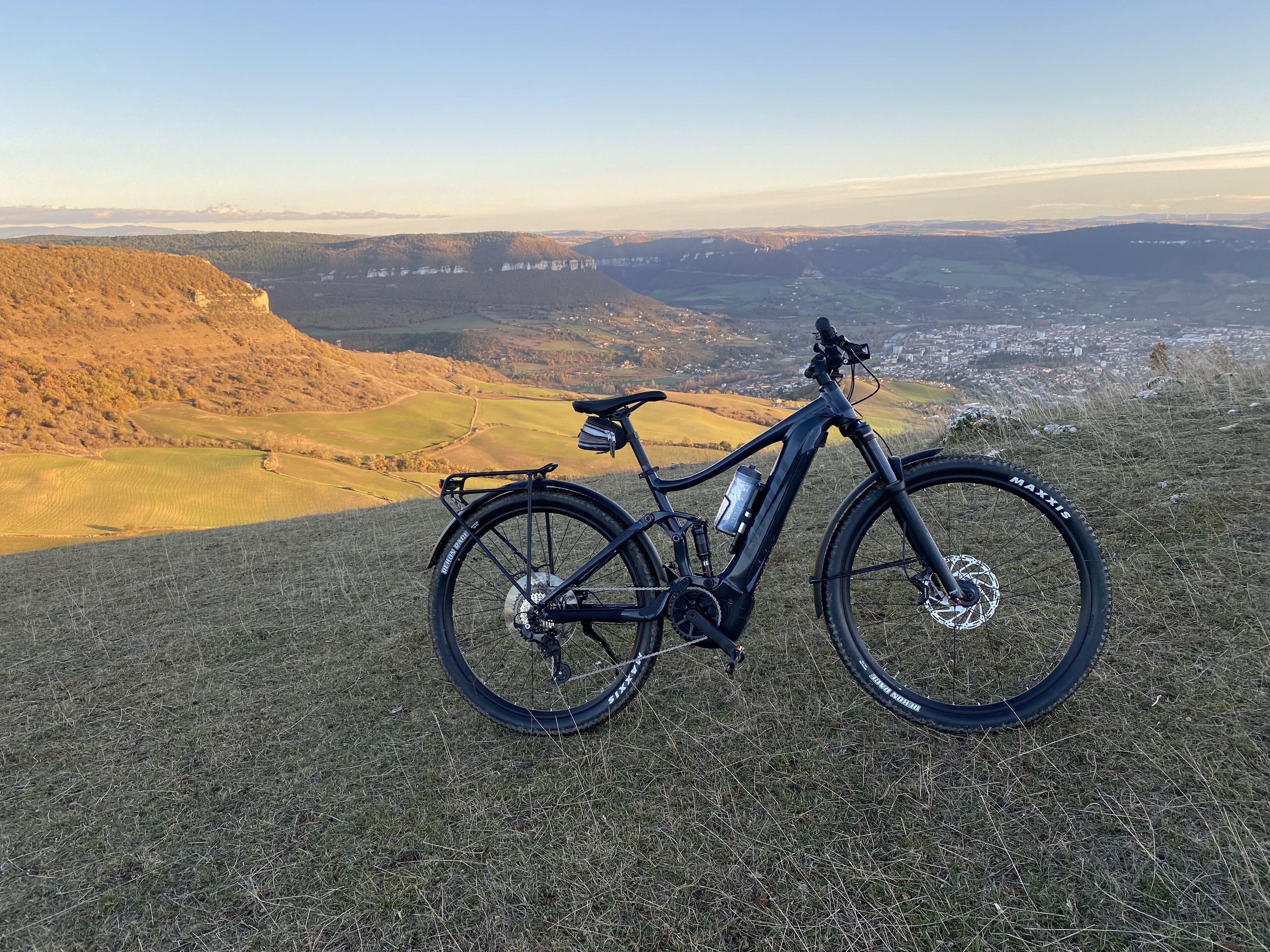 A black mountain bike with a water bottle and a rear rack, parked on a grassy hill overlooking a valley with fields and distant hills.