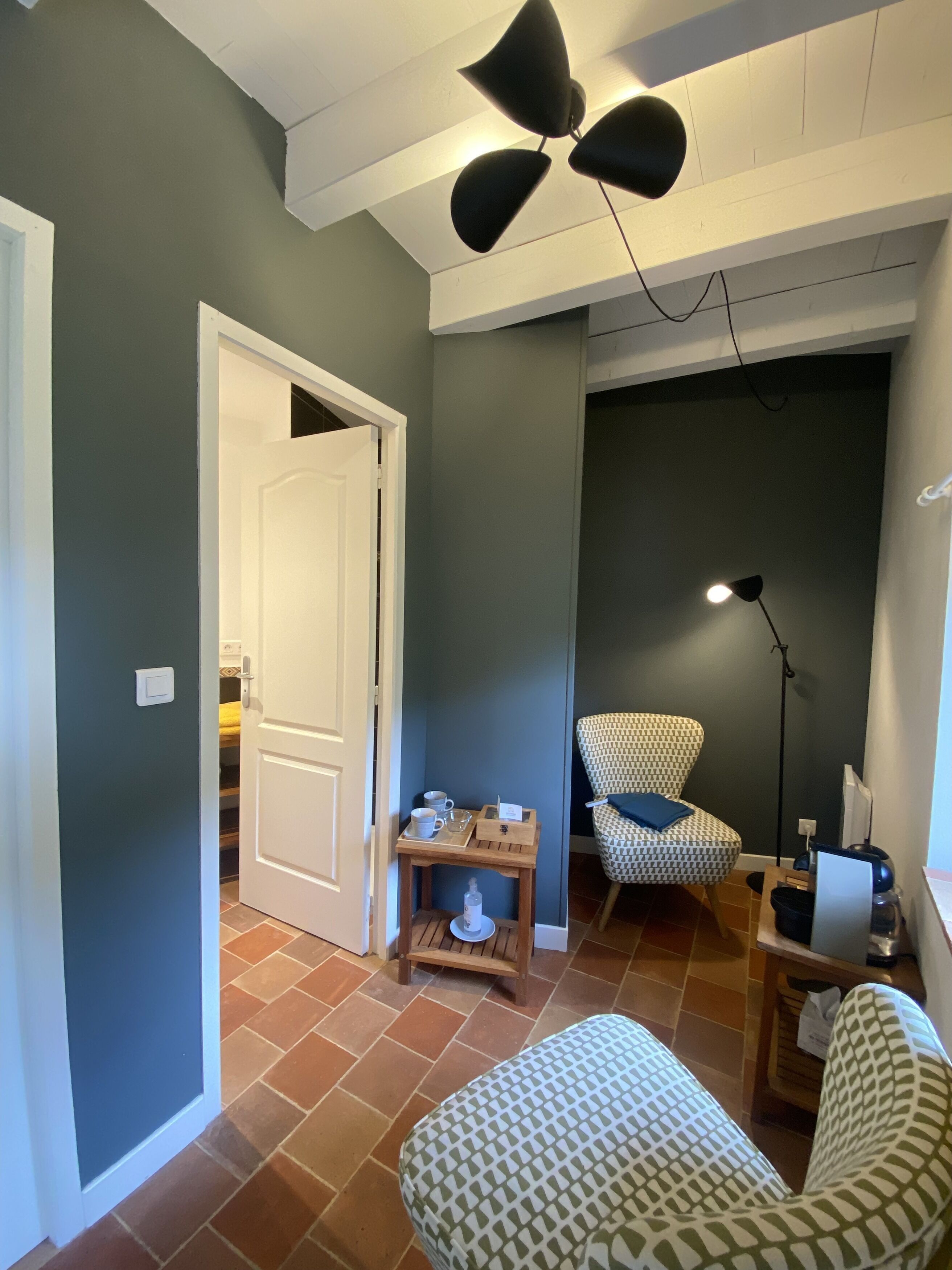Una habitación con un sillón estampado, una pequeña mesa de madera con una bandeja, una silla y un escritorio con una computadora.