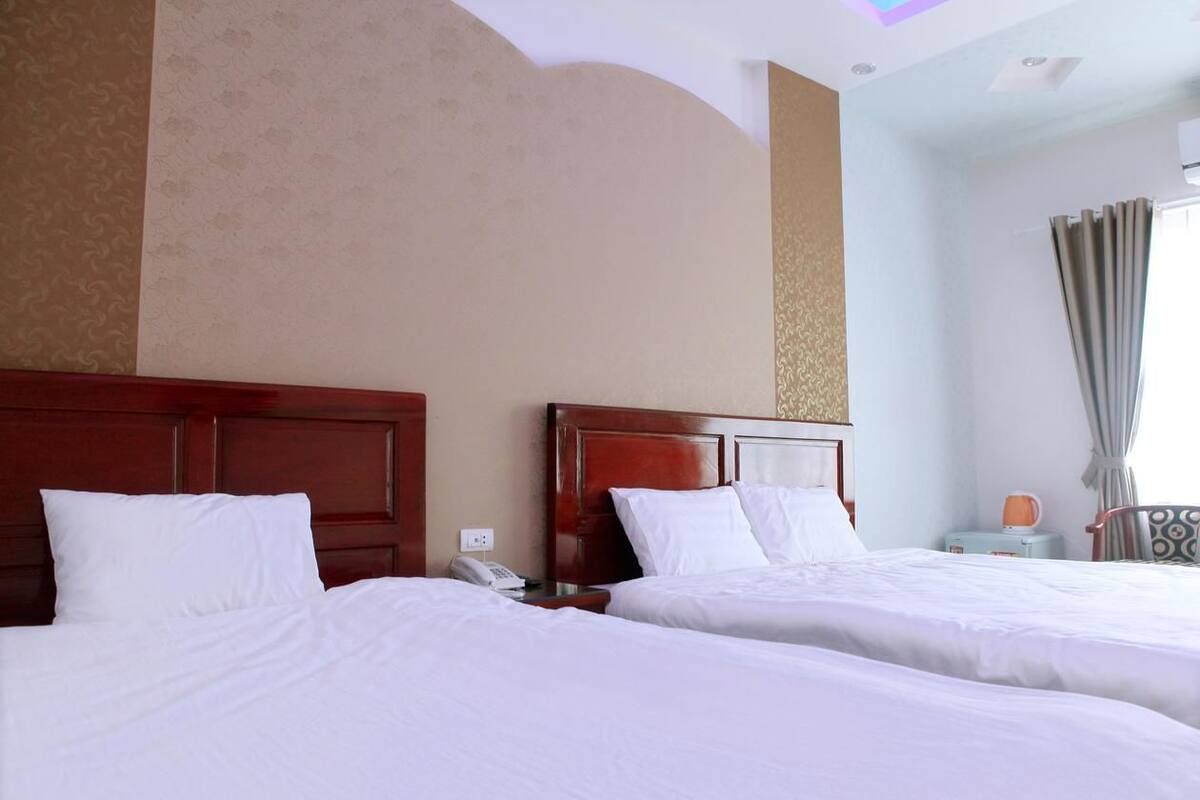 Quarto de hotel com duas camas, cabeceiras de madeira, mesa de cabeceira com telefone e uma cadeira com almofada estampada.