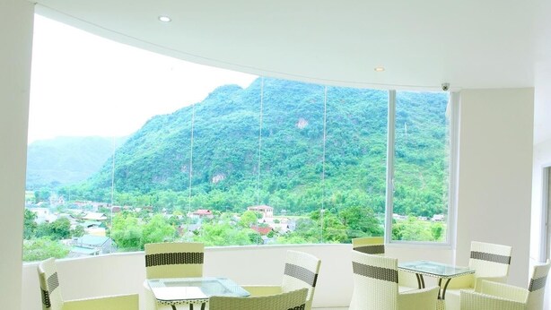 Una habitación con un ventanal que ofrece vistas a un paisaje montañoso y un pueblo en la ladera. Hay sillas de mimbre y una pequeña mesa en la habitación.