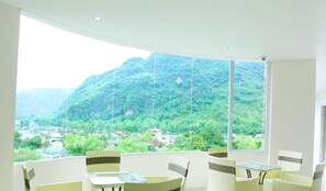 A room with a large window offering a view of a mountainous landscape and a village below. There are wicker chairs and a small table in the room. - Ngoc Bach Hotel (Mai Chau)