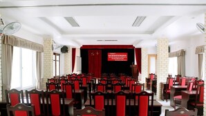 Un salón de conferencias con filas de sillas y mesas de madera, un escenario con cortina roja y una pared de ladrillo.