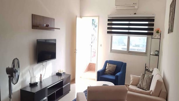 A living room with a flat-screen TV, a sofa, a blue armchair, a small table, and a standing fan. - Résidence Lumière (Abidjan)