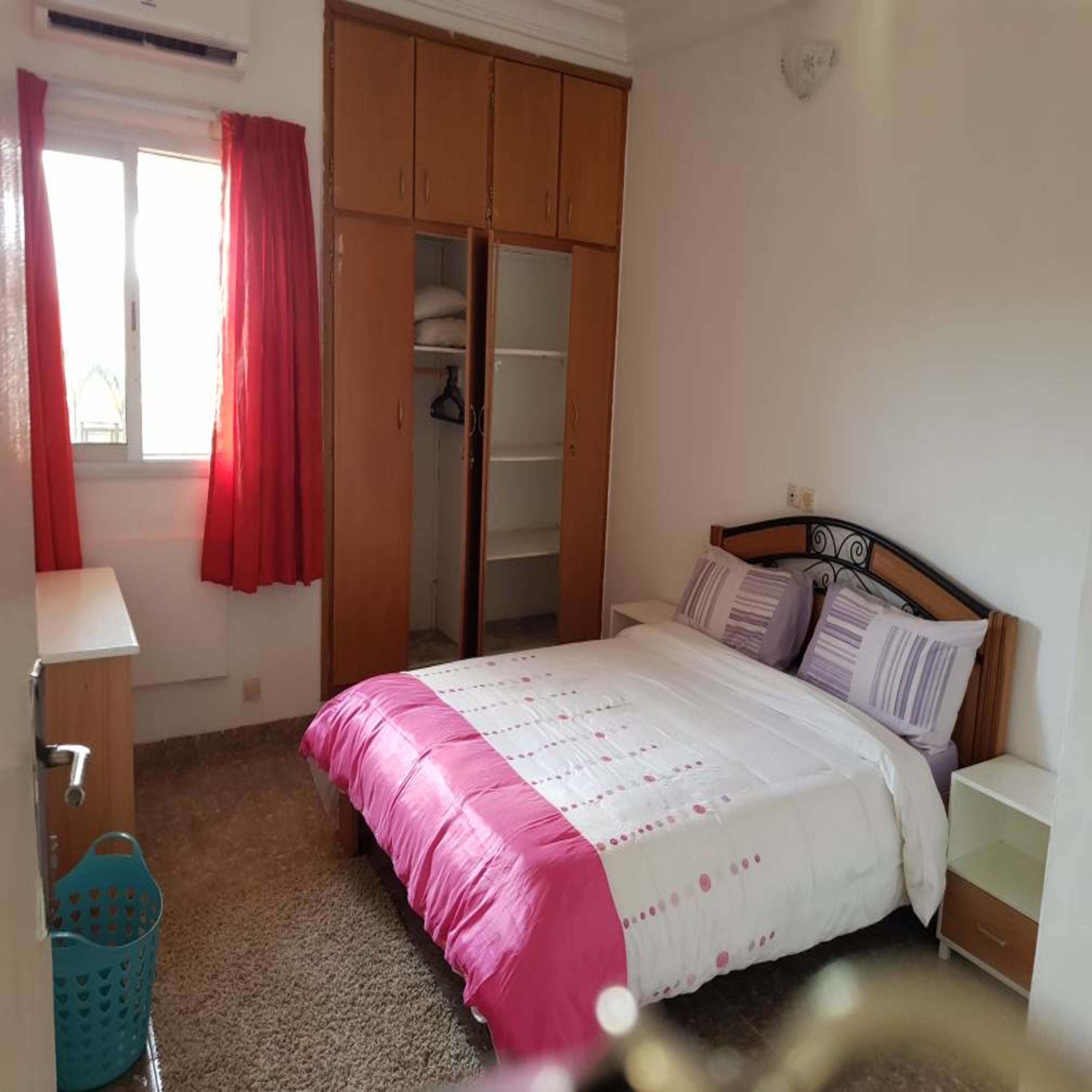 A bedroom with a bed, a wardrobe, a window with curtains, and a small bedside table.