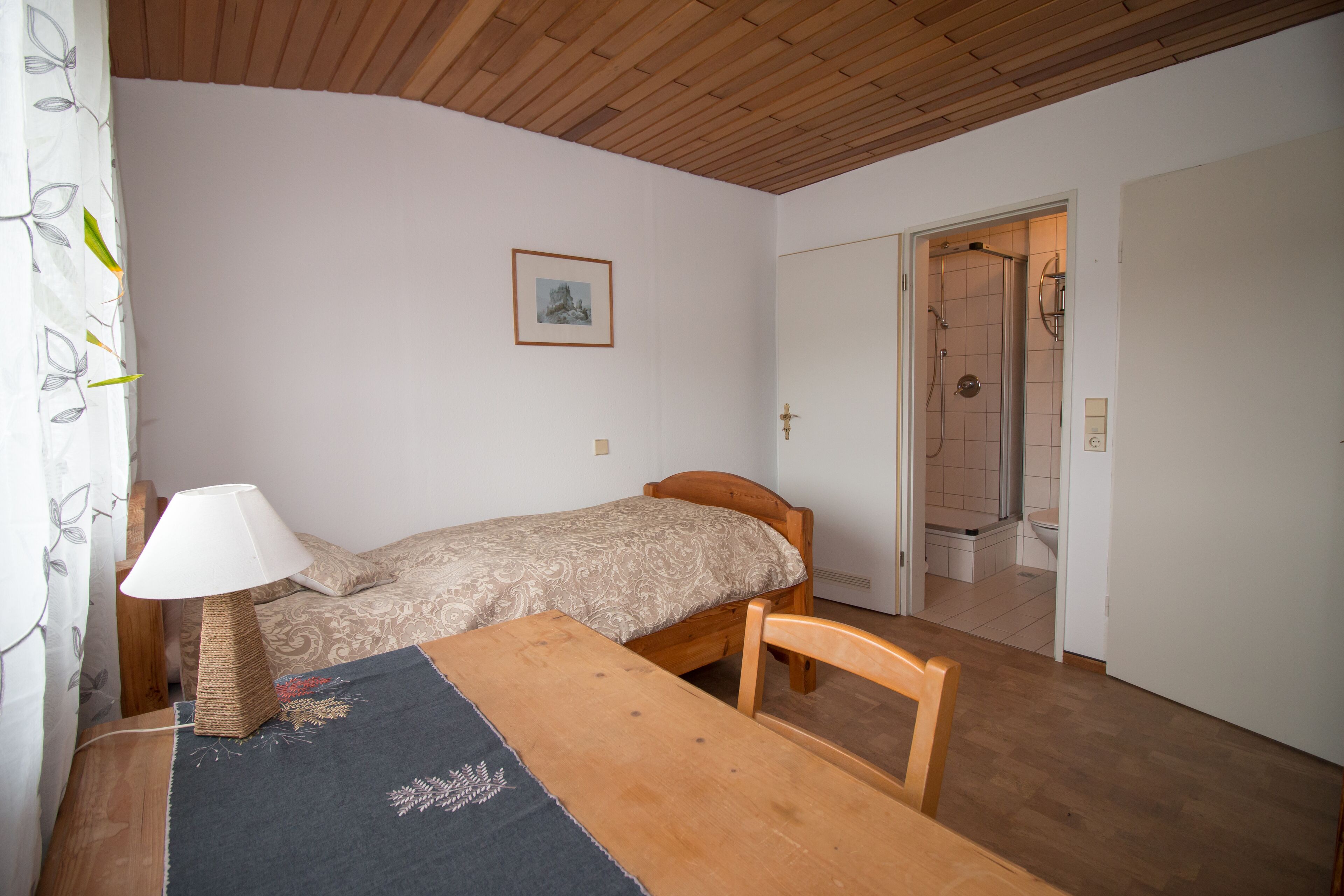 Ein Schlafzimmer mit einem Einzelbett, einem Holztisch, einer Lampe und einem gerahmten Bild an der Wand.