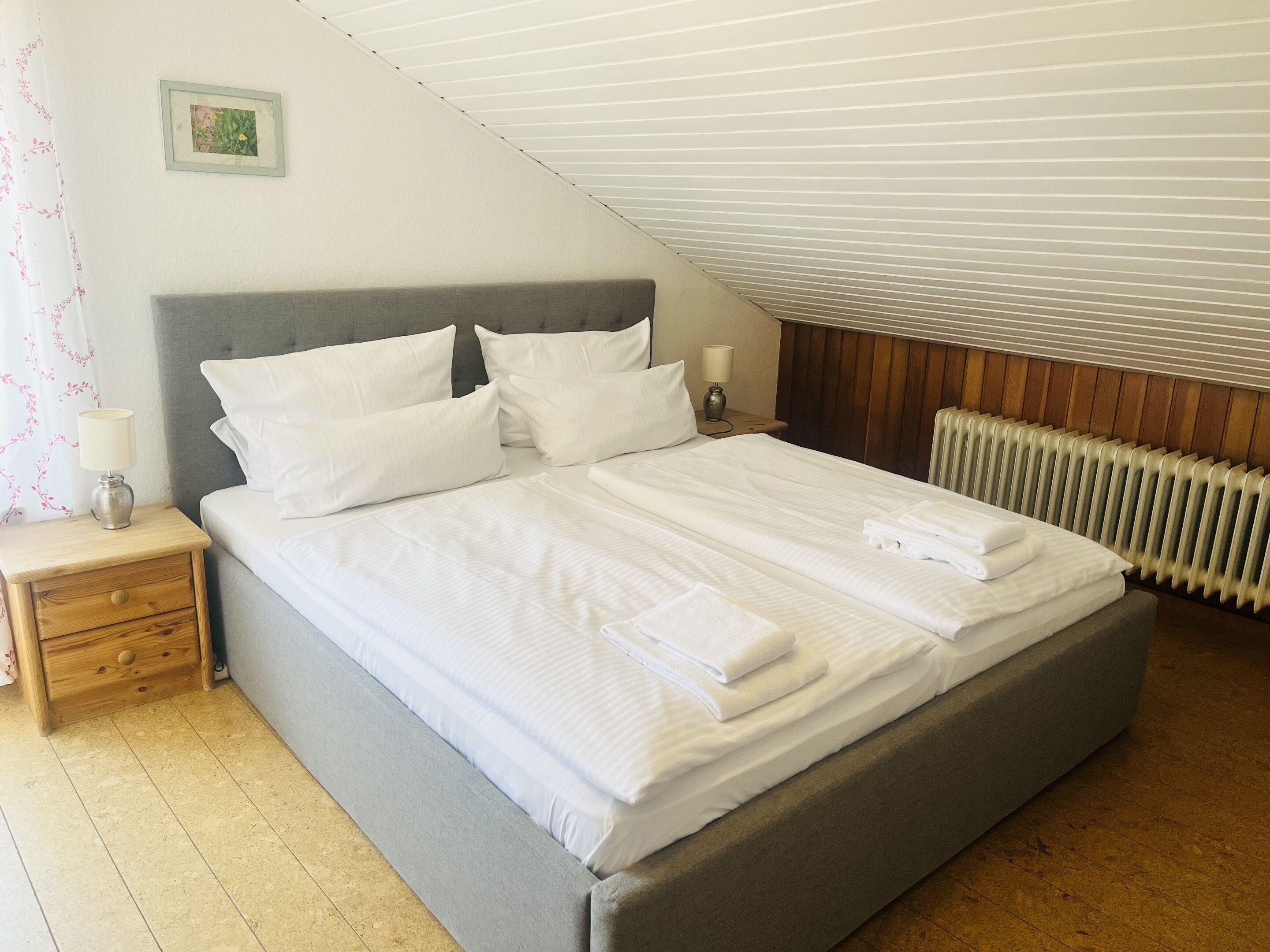 A neatly made bed with pillows and folded towels, a bedside table with a lamp, and a radiator on the wall.
