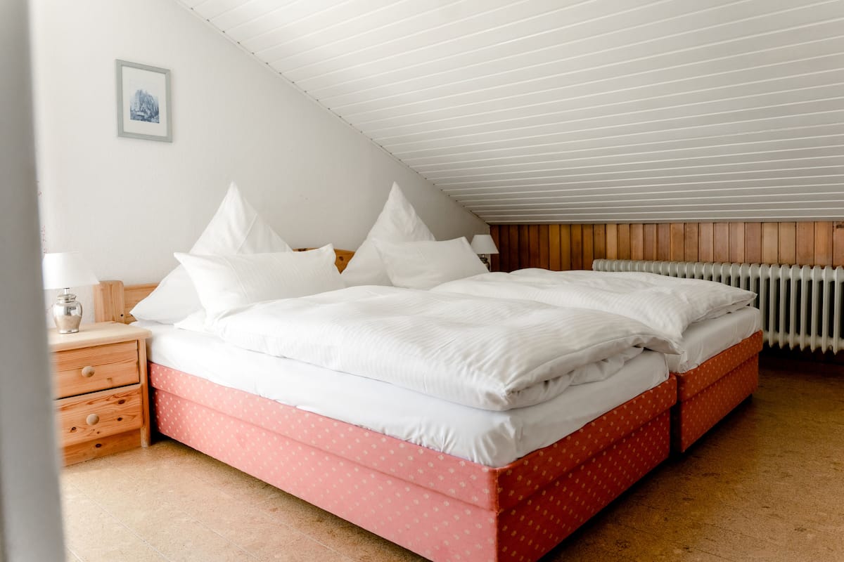 Un dormitorio con techo inclinado, cama con ropa de cama blanca, cabecera de madera, mesita de noche con lámpara y un radiador.