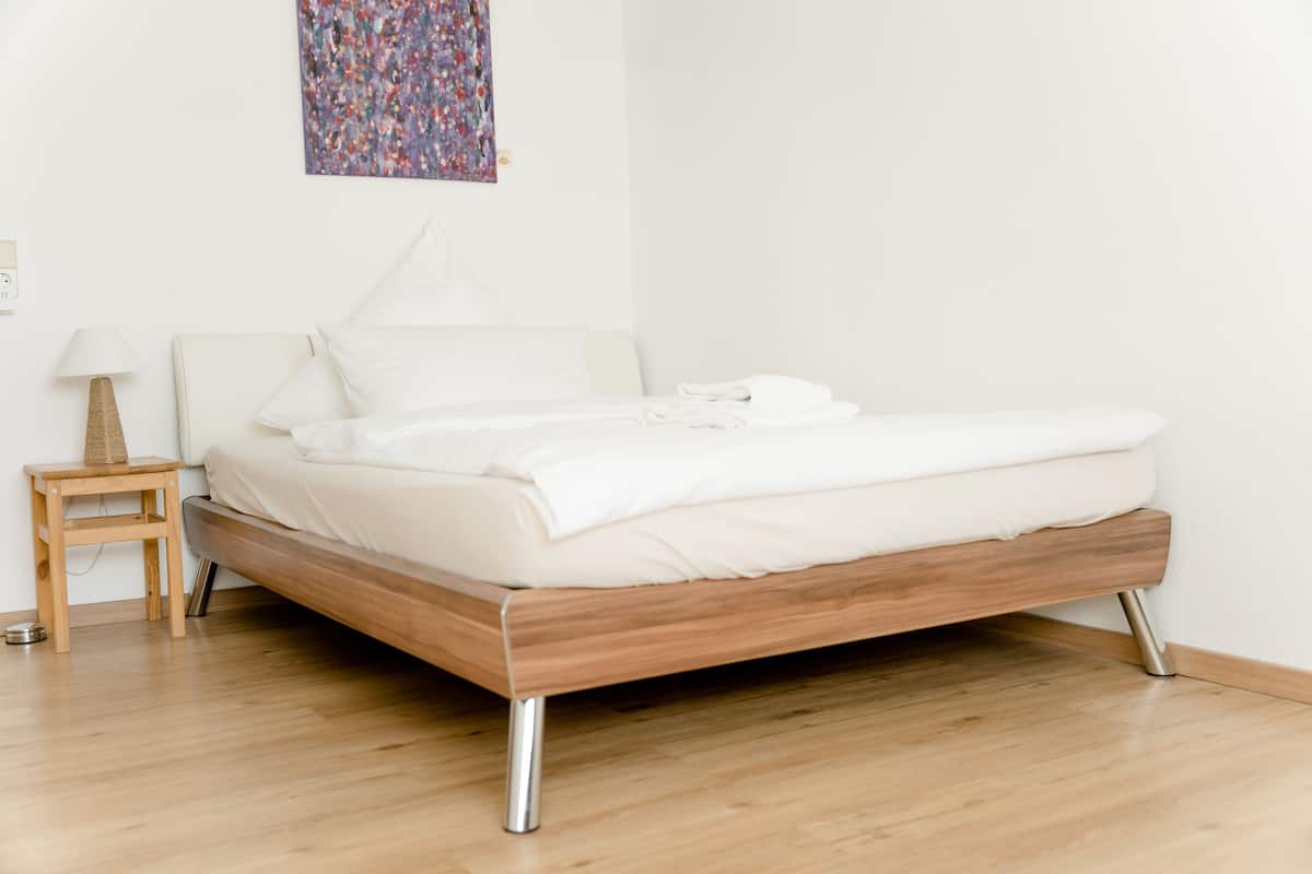 Una cama bien hecha con sábanas blancas en una habitación con suelo de madera y una mesita de noche de madera con una lámpara.