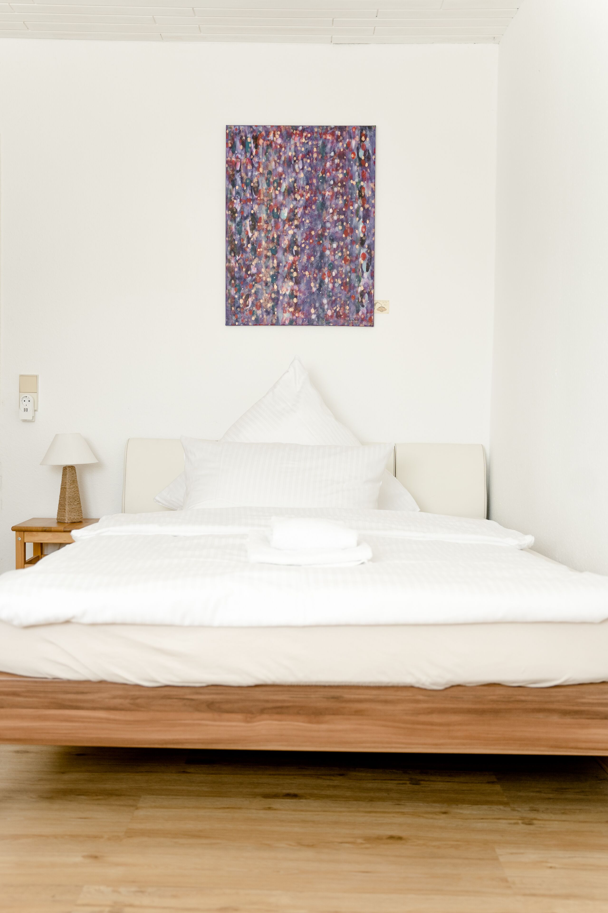 A minimalist bedroom with a wooden bed frame, white bedding, and a colorful abstract painting on the wall.