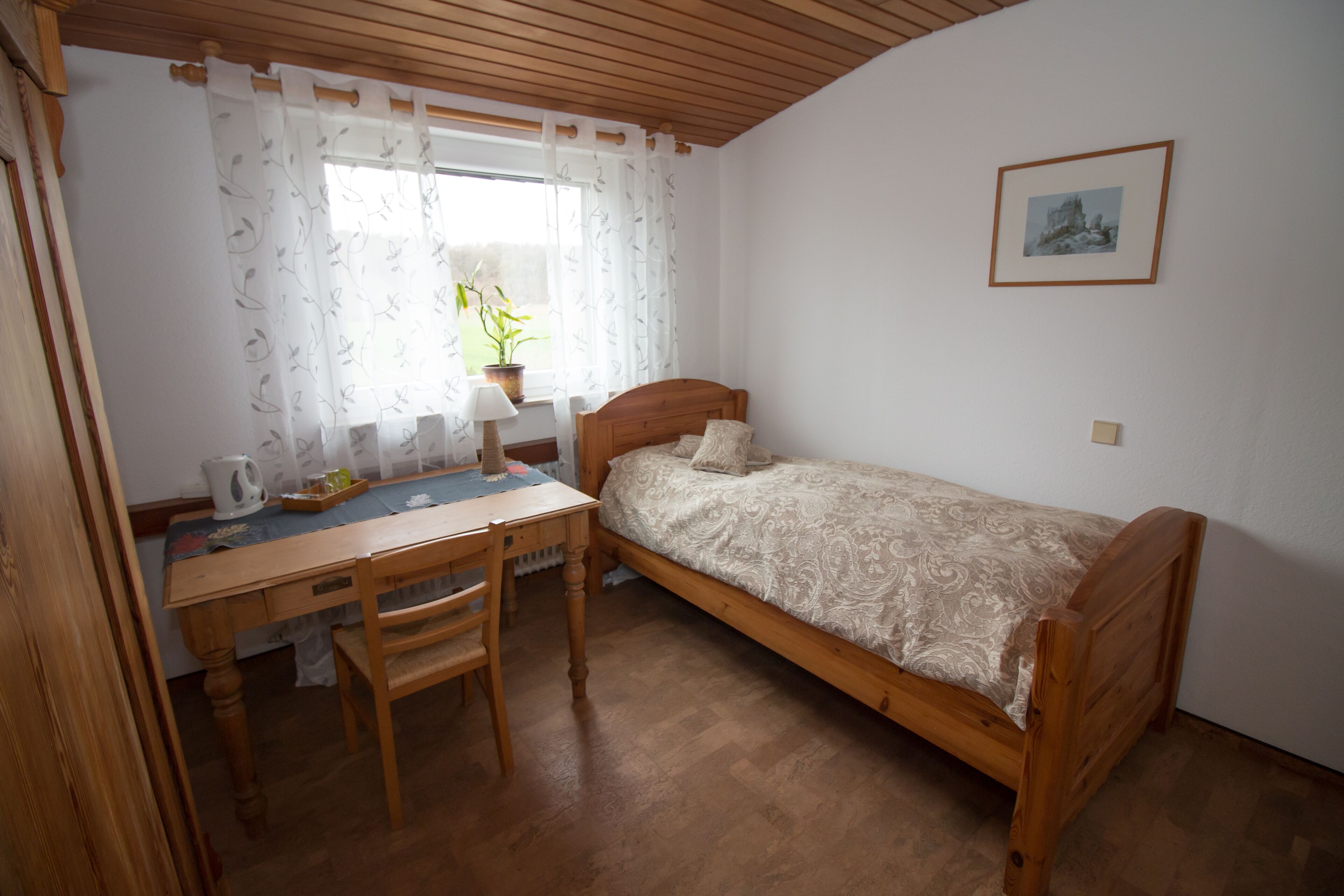 Une petite chambre avec un lit simple, un bureau et une chaise en bois, une fenêtre avec des rideaux légers, une plante en pot et un tableau encadré au mur.