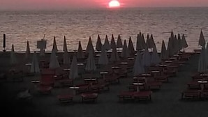 A beach with rows of umbrellas and tables, a sunset in the background, and a calm sea.
