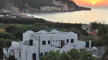 Villa blanca llamada 'Albergc Villa Amare' con balcón, rodeada de vegetación y con vistas a una zona costera con acantilados y el mar.