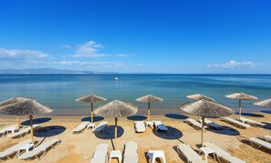 A beach with sun loungers and thatched umbrellas, a clear blue sea, and a boat in the distance. - Elpiniki Luxury Apartments (Nea Propontida)