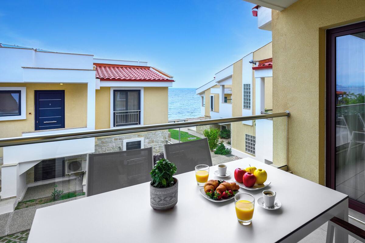 Un balcon avec une table préparée pour le petit-déjeuner, comprenant des fruits, du jus et du café, donnant sur une zone côtière avec des bâtiments et la mer.